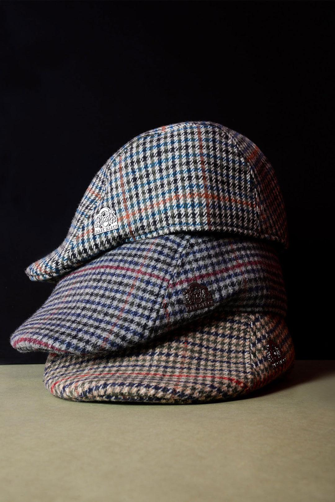 Three plaid flat caps in different color patterns are stacked on top of each other against a dark background.
