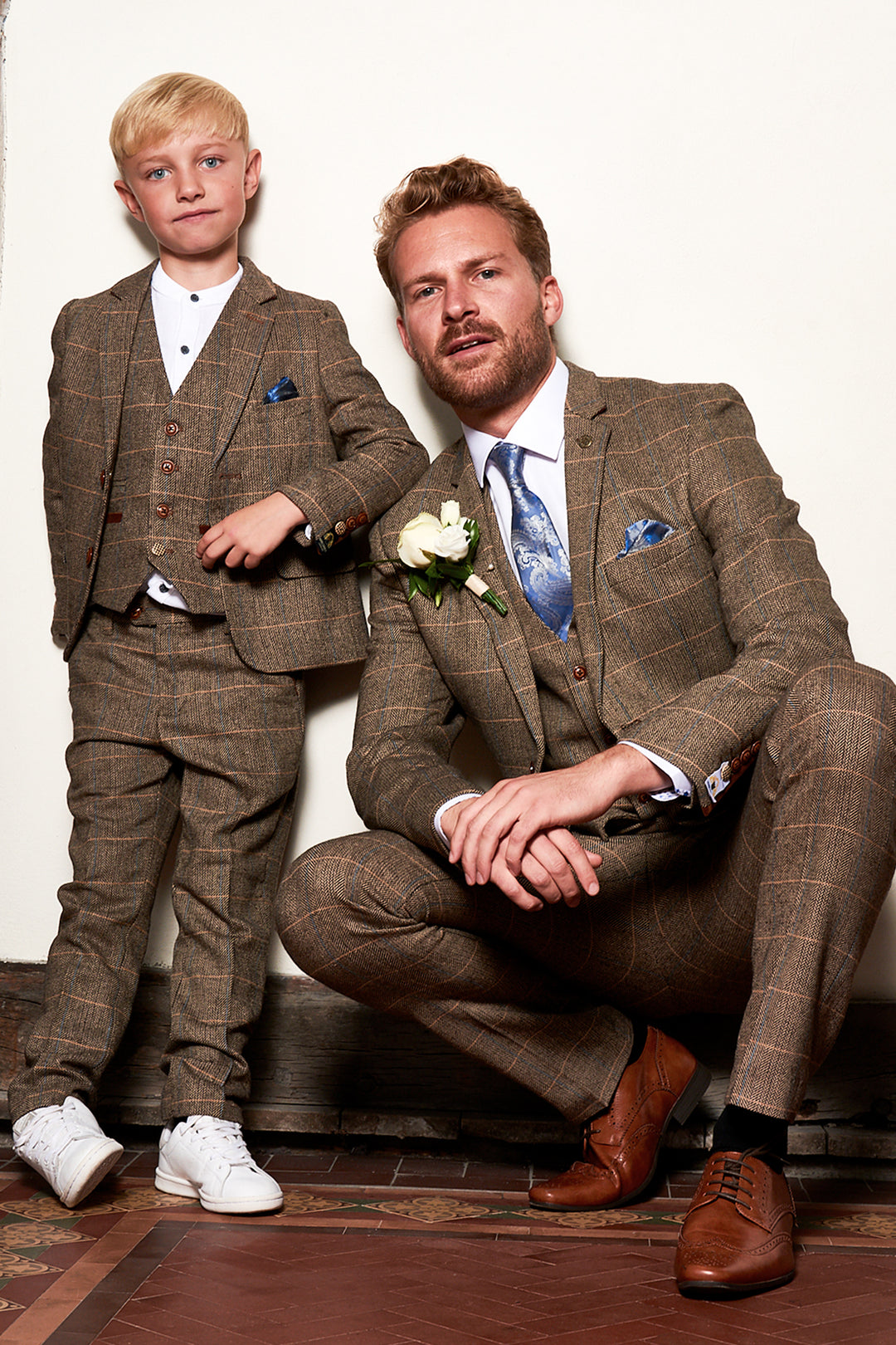 A man and a young boy in matching brown plaid suits pose together; the man is squatting with one knee down, and the boy stands beside him. The boy wears white sneakers, and the man wears brown dress shoes.