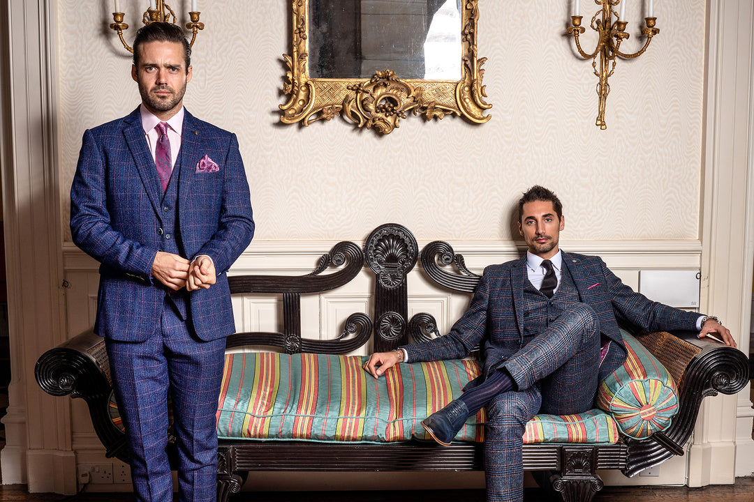 Two men in patterned suits pose in an elegant room; one stands beside a vintage striped sofa while the other sits on it. Ornate gold-framed mirror and wall sconces decorate the background.