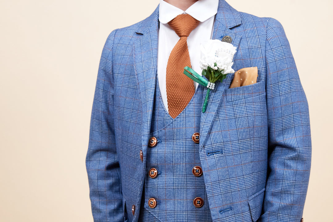 Man wearing a blue plaid suit with a brown tie and pocket square, white dress shirt, and a white flower boutonniere on the lapel, standing against a light beige background.