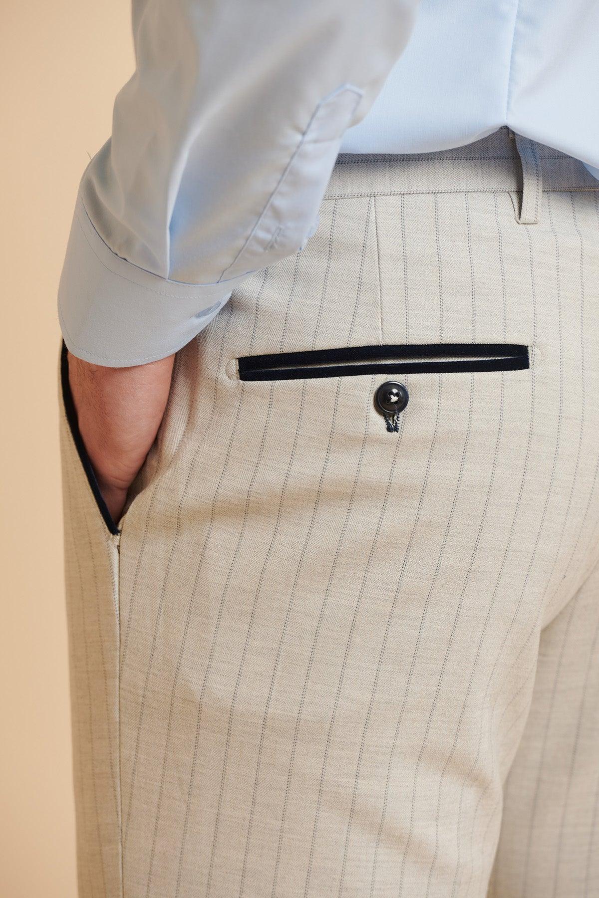 A man’s left hand is in the pocket of tan pinstripe pants from The Everton Collection | Beto In GRANT Stone Pinstripe Double Breasted Suit by Marc Darcy. A light blue shirt is visible at the top, completing the stylish three-piece look.