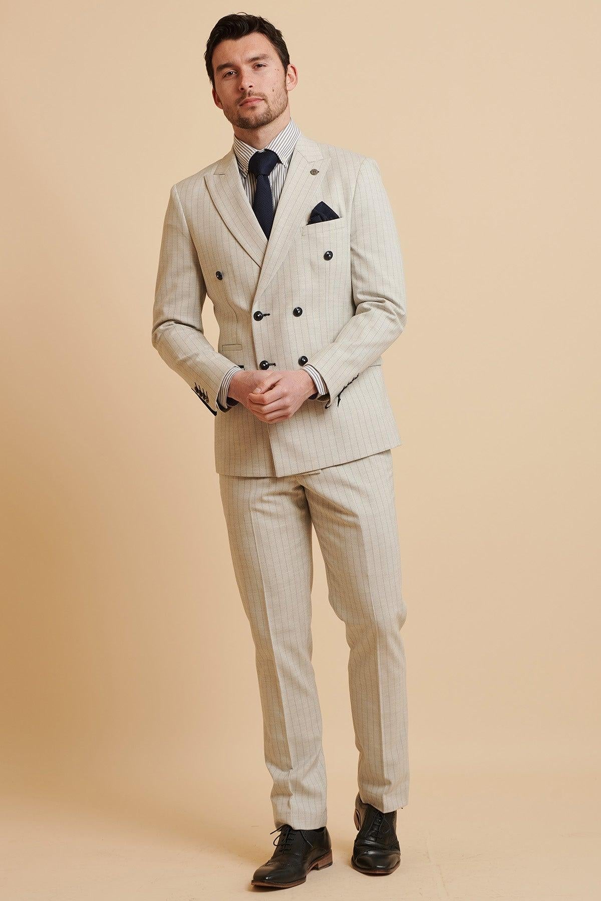 A man wears The Everton Collection | Beto In GRANT Stone Pinstripe Double Breasted Suit by Marc Darcy with a navy tie and black dress shoes, standing against a tan background.