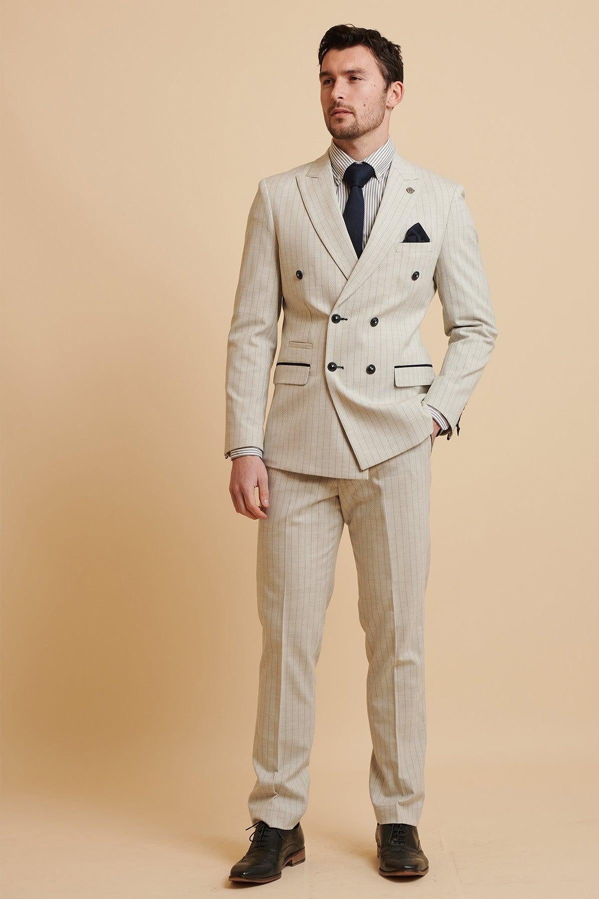 A man models The Everton Collection | Beto In GRANT Stone Pinstripe Double Breasted Suit by Marc Darcy, styled with a blue tie, striped white shirt, and black dress shoes, standing against a plain beige background.