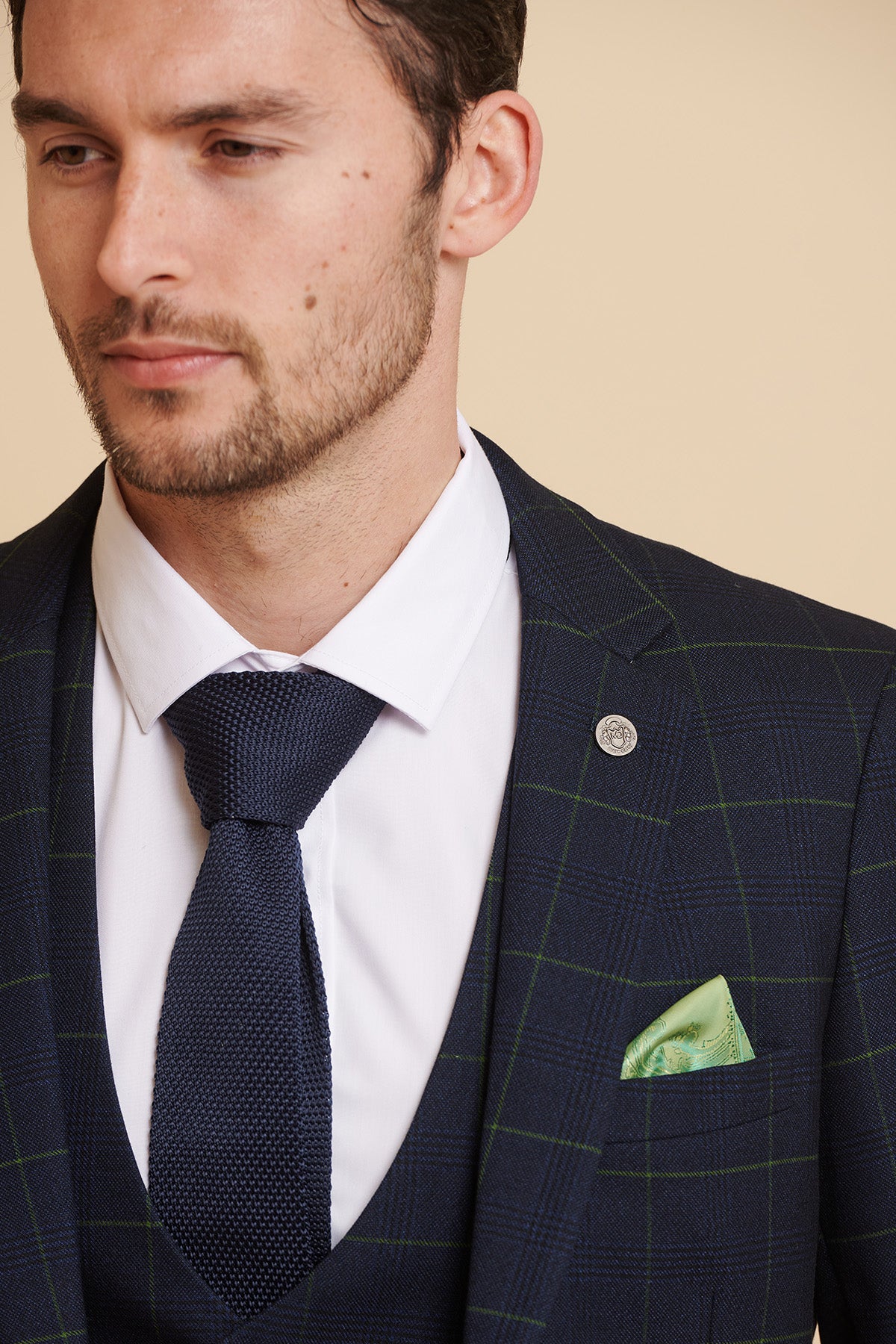 A man wears Marc Darcy's The Everton Collection Beto In EDINSON Navy Green Check Two Piece Suit with a white shirt, navy tie, green paisley pocket square, and lapel pin.