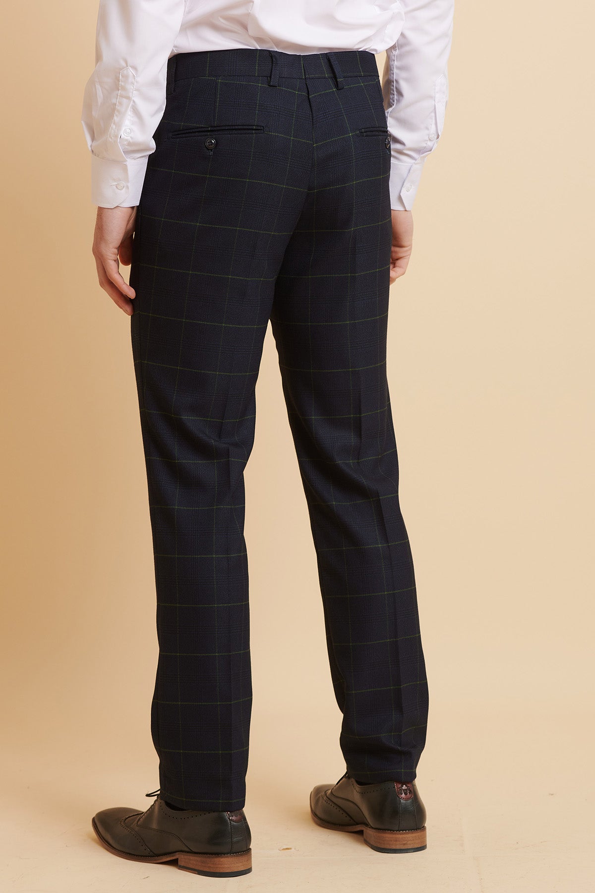 A person in The Everton Collection Beto In EDINSON Navy Green Check Two Piece Suit by Marc Darcy stands facing away. Slim tailored fit pants and dark dress shoes complete the classic look against a beige background.