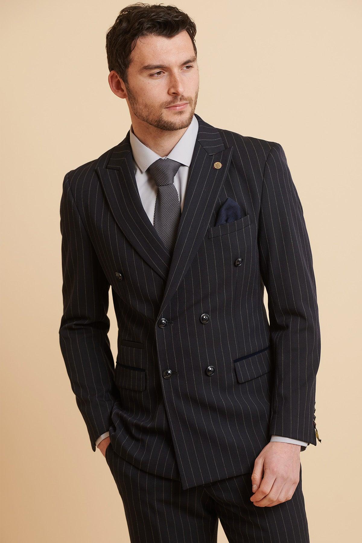 A man wears the Marc Darcy ROCCO Navy Pinstripe Double Breasted Blazer with a white shirt and gray tie, standing against a beige background, one hand in his pocket and looking slightly to the side.