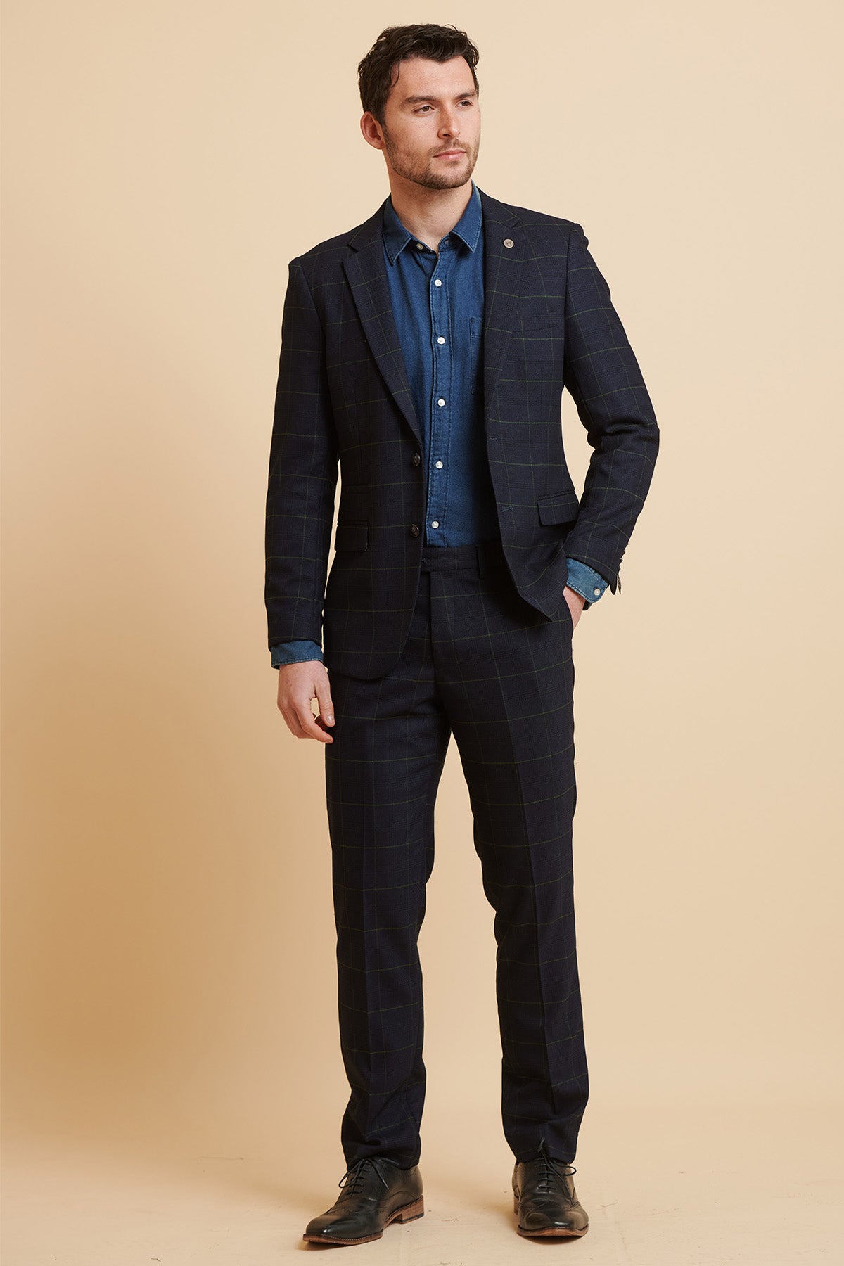 A man stands against a beige background in the Marc Darcy Everton Collection Beto In EDINSON Navy Green Check Two Piece Suit, paired with a blue shirt and black dress shoes, looking slightly to the side with a neutral expression.
