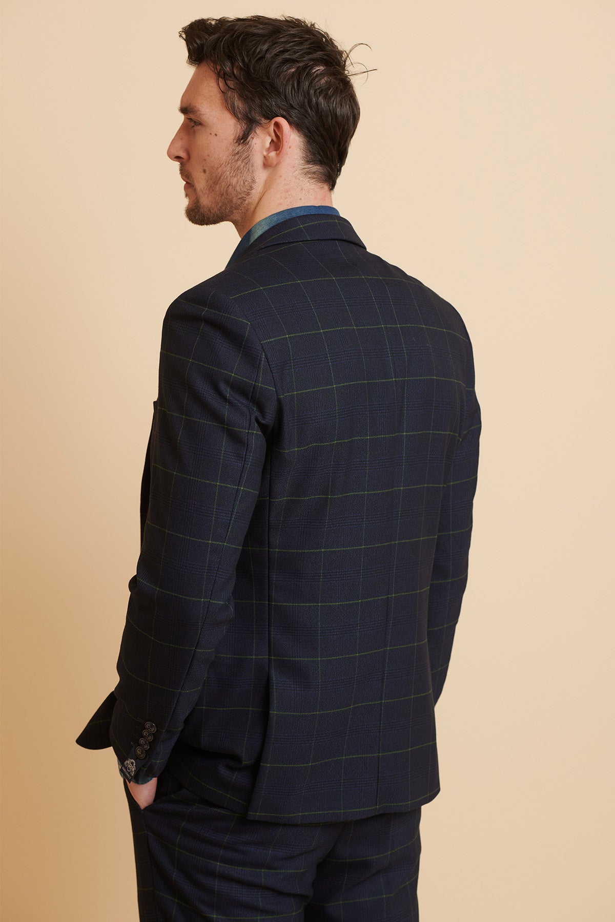 A man with short dark hair, wearing the Marc Darcy Edinson Navy Green Check Blazer from The Everton Collection, stands with his back to the camera and one hand in his pocket against a plain beige background.