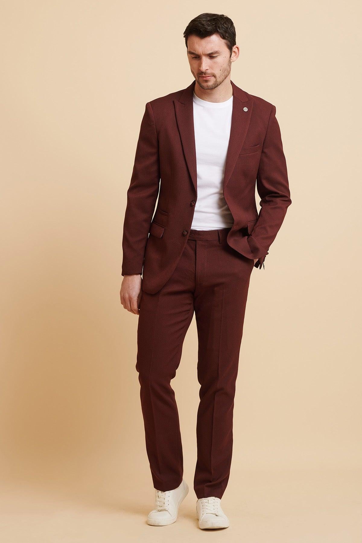 A man poses against a beige background wearing the marcdarcy MAX - Wine Two Piece Suit with a white t-shirt and sneakers, his slim fit blazer open, one hand in his pocket and a relaxed expression.