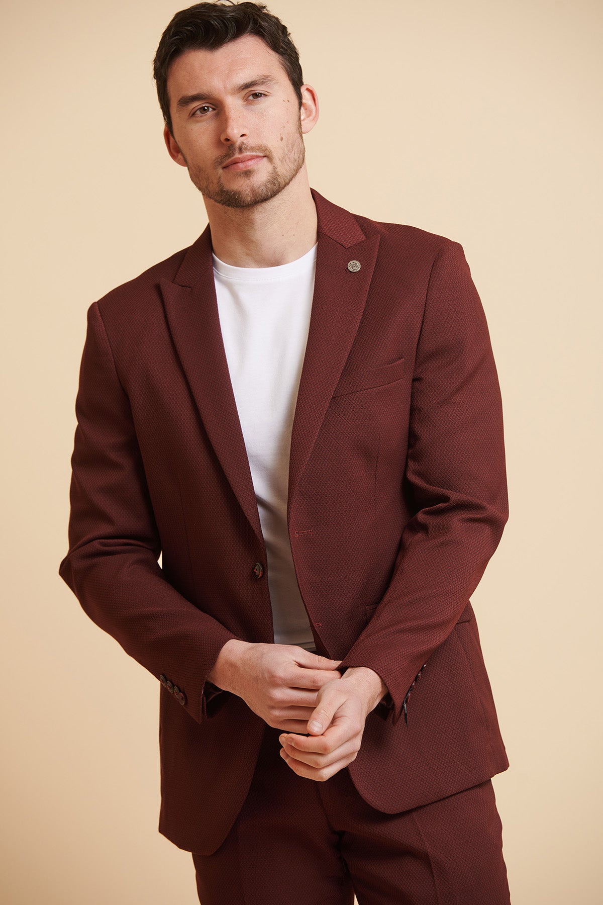 A man stands against a tan background with his hands clasped, wearing the marcdarcy MAX Wine Tailored Blazer over a white t-shirt.