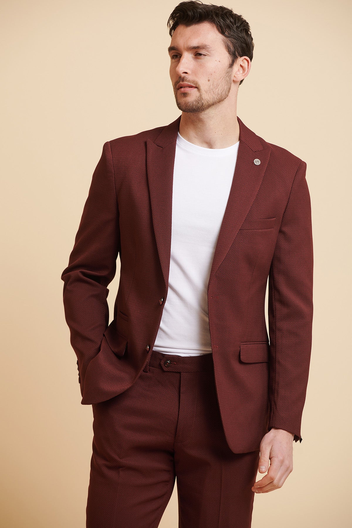 A man models the marcdarcy MAX - Wine Tailored Blazer over a white t-shirt, standing against a beige background and gazing to the side with one hand in his pocket.