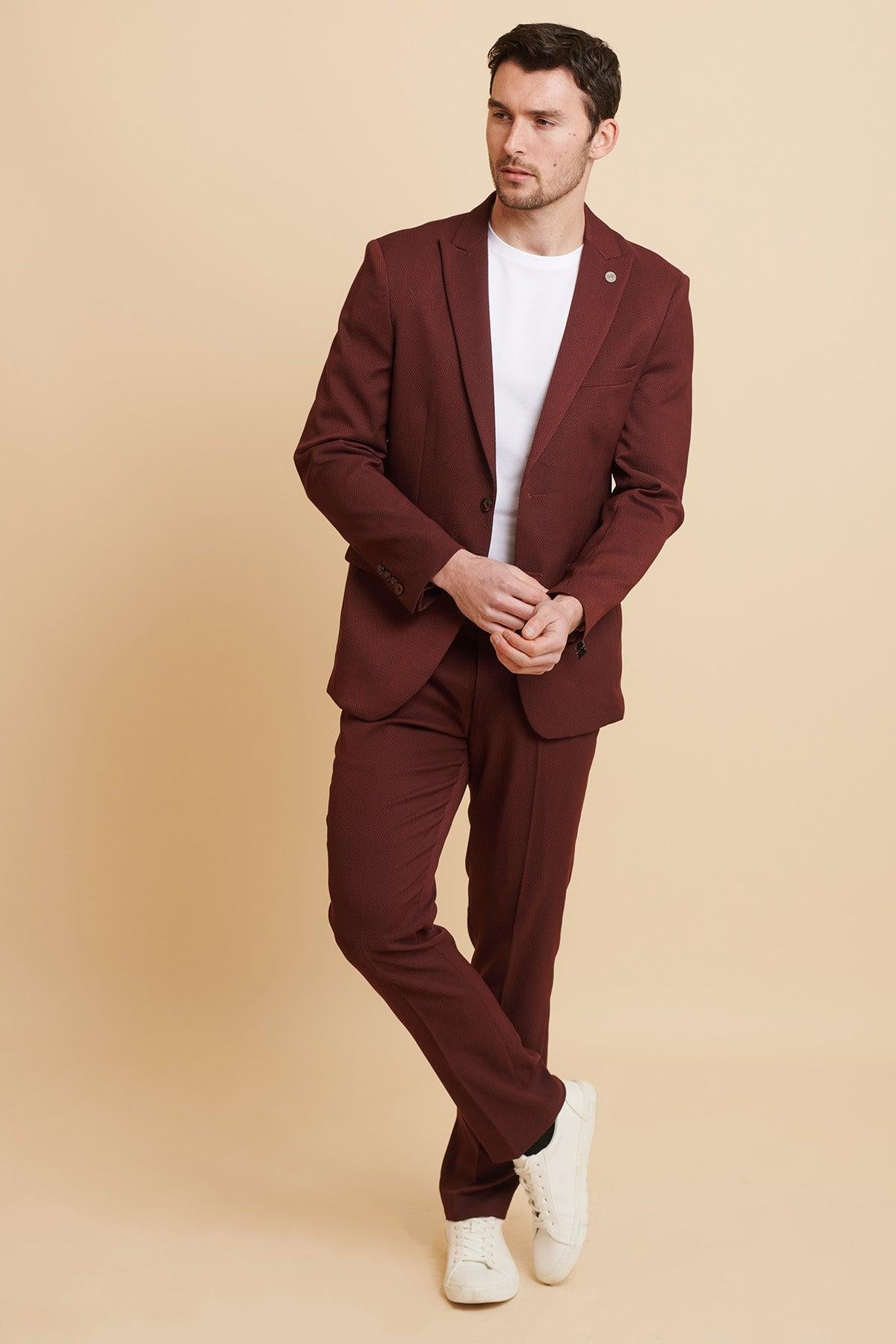 A man stands against a beige background wearing the marcdarcy MAX Wine Two Piece Suit with a white t-shirt and sneakers. He crosses his legs, clasps his hands, glances sideways, and adds flair with a camouflage pocket square.