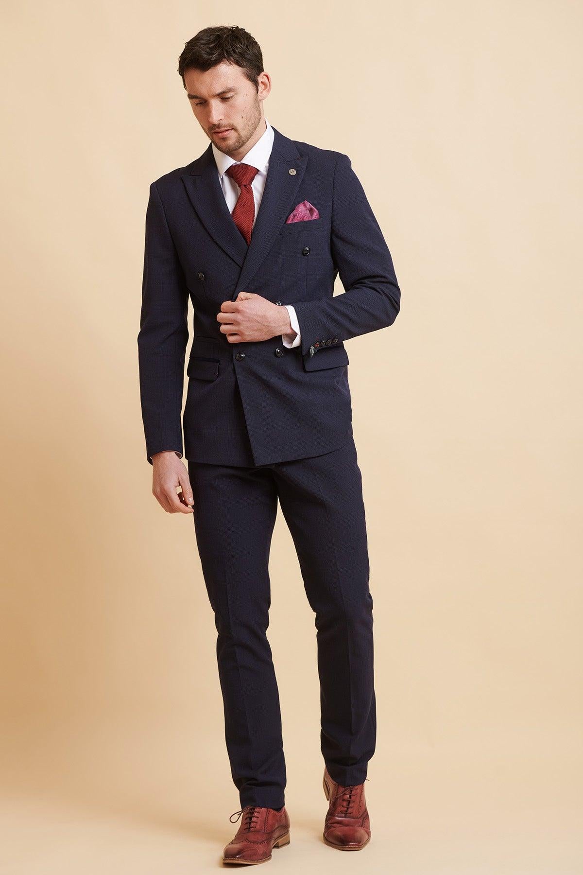 A man in the Official West Ham United Club Suit—BROMLEY Navy Double Breasted Suit by Marc Darcy—paired with a white shirt, red tie, burgundy pocket square, and brown wingtip shoes stands against a tan background.