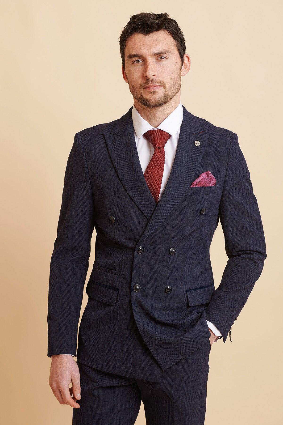 A man poses with a hand in his pocket against a tan background, wearing the Marc Darcy Official West Ham United Club Suit | BROMLEY Navy Double Breasted Suit, paired with a white shirt and red tie.