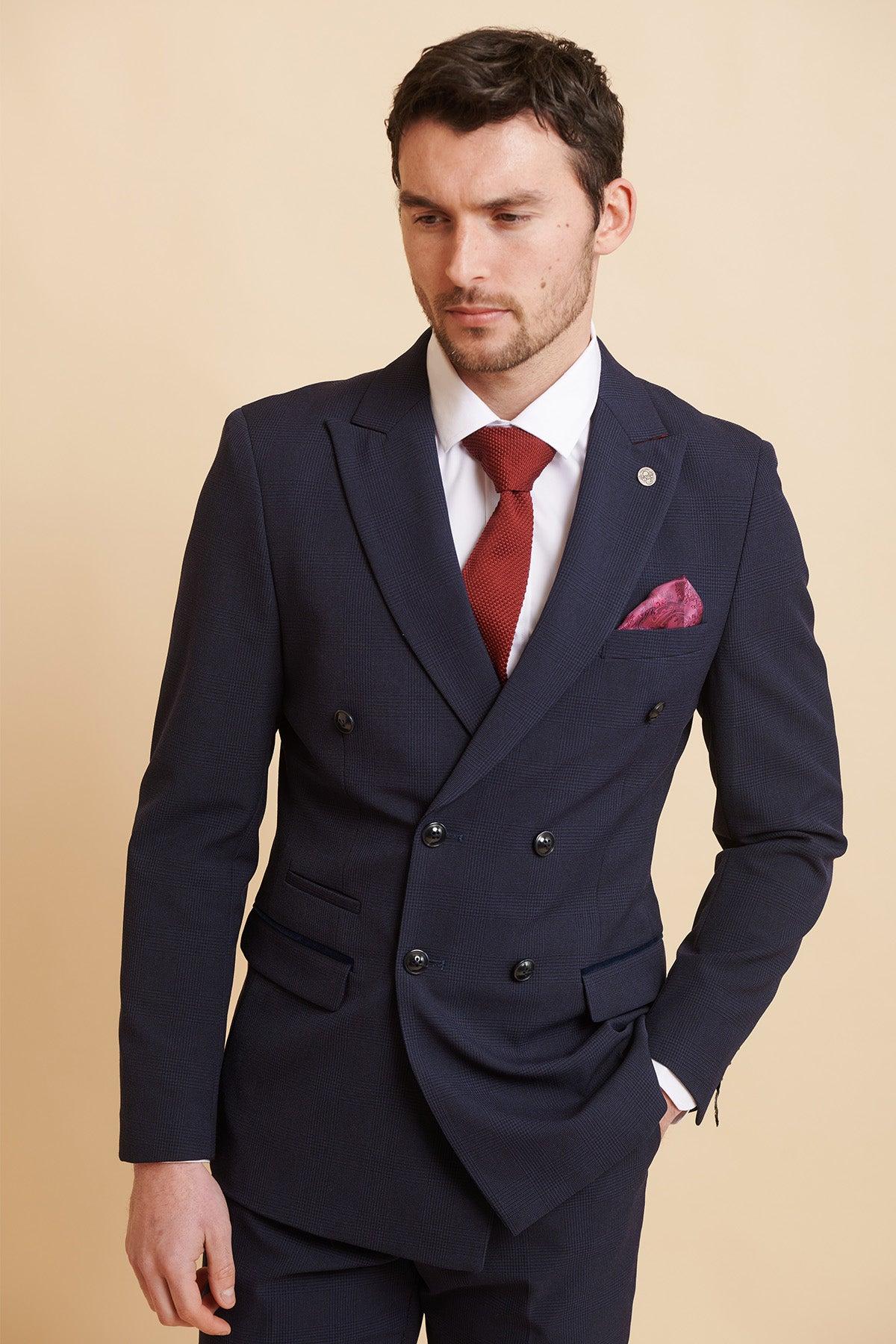 A man wears the Official West Ham United Club Suit | BROMLEY Navy Double Breasted Suit by Marc Darcy, paired with a white shirt, red tie, and pink pocket square, with his hand in his pocket.