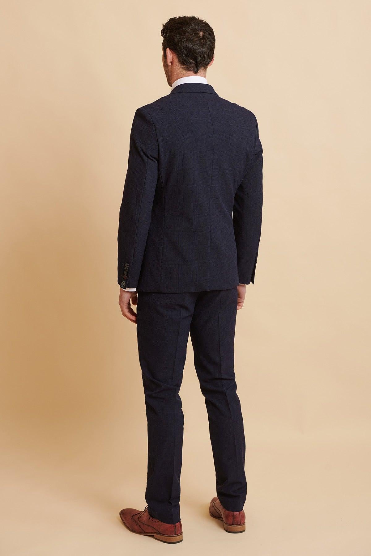 Back view of a man wearing the Official West Ham United Club Suit, the BROMLEY Navy Double Breasted Suit by Marc Darcy, paired with a white shirt and brown dress shoes against a cream background.