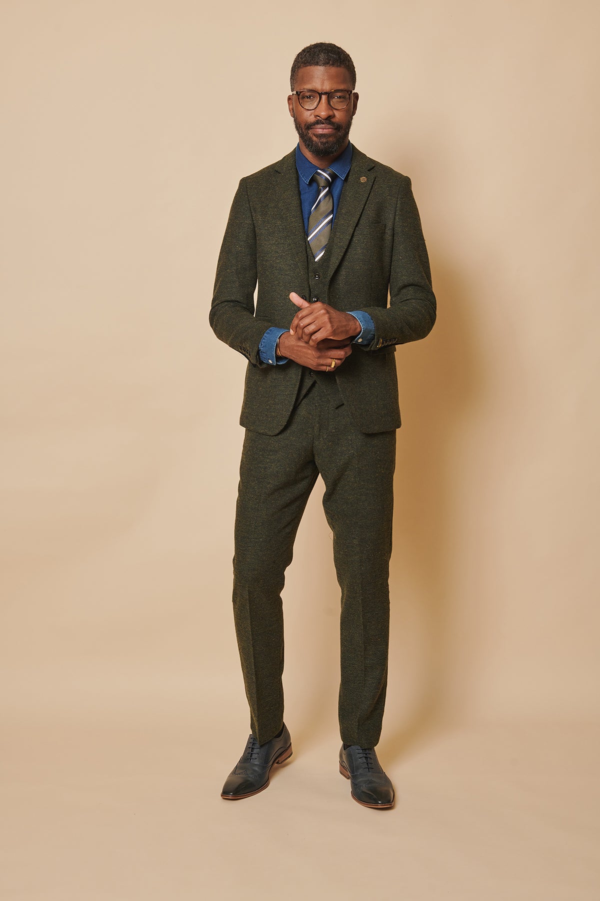 A man stands against a beige background wearing the marcdarcy MARLOW Olive Green Tweed Three Piece Suit, a blue shirt, striped tie, and dress shoes. He has short hair, glasses, and looks forward with his hands clasped.