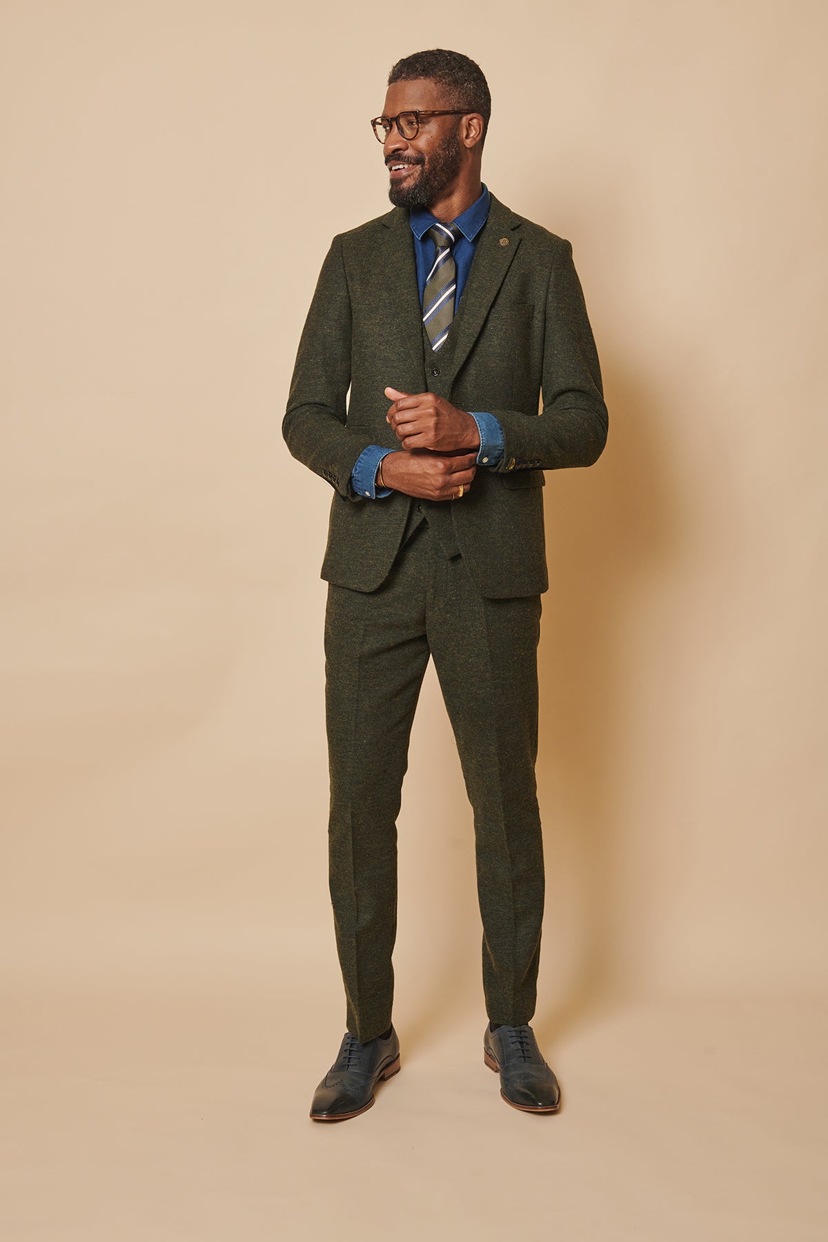 A man wearing glasses and the marcdarcy MARLOW Olive Green Tweed Three Piece Suit with a blue shirt, patterned tie, and dark shoes stands smiling against a beige background.
