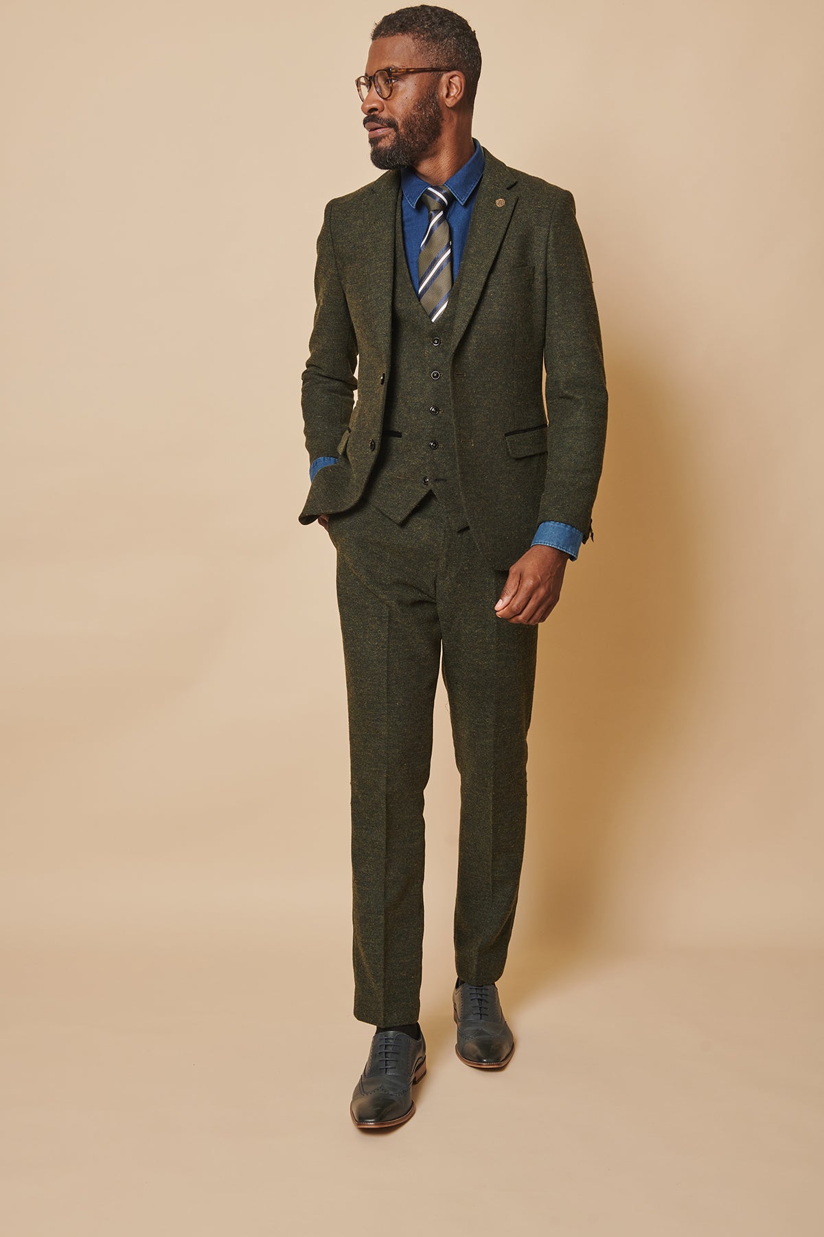 A man in the marcdarcy MARLOW olive green tweed three piece suit, blue dress shirt, striped tie, and dark shoes stands against a beige background with one hand in his pocket, looking to the side.