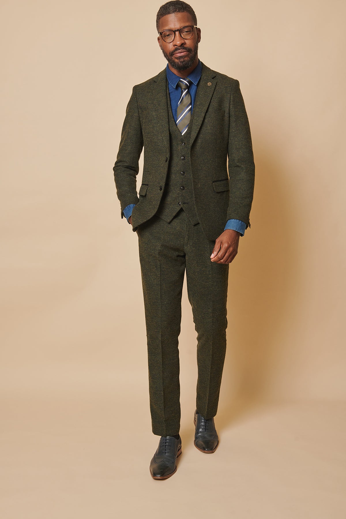 A man poses confidently against a beige backdrop, wearing the marcdarcy MARLOW Olive Green Tweed Three Piece Suit in a slim fit, paired with a blue shirt, striped tie, black shoes, glasses, and sporting short hair and a beard. One hand rests in his pocket.