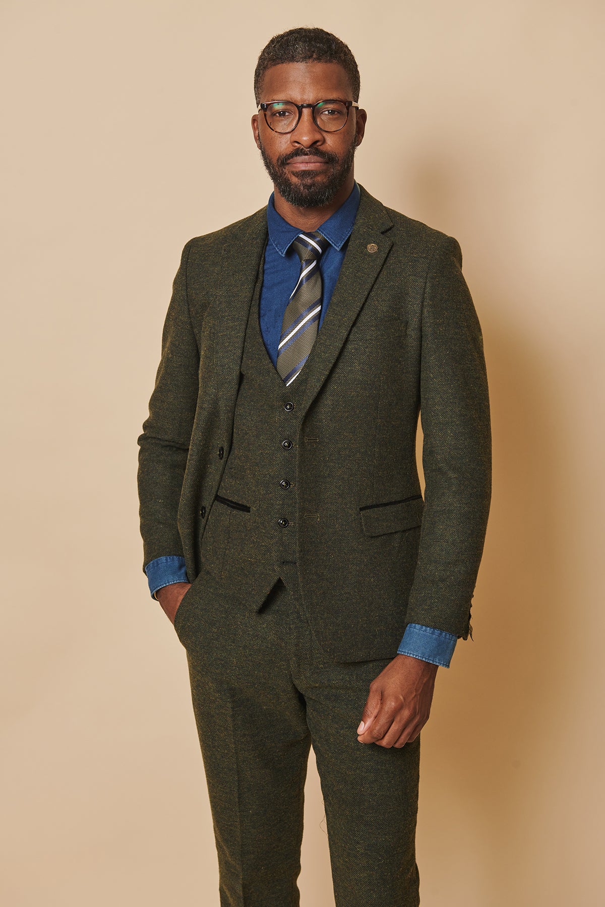 A man with short hair, a beard, and glasses stands against a beige background wearing the marcdarcy MARLOW Olive Green Tweed Blazer (slim fit wool-poly mix), with a blue dress shirt, striped tie, vest, and one hand in his pocket.