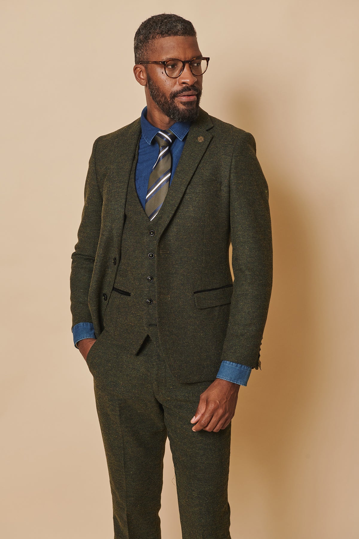 A man with short hair, a beard, and glasses wears the marcdarcy MARLOW Olive Green Tweed Blazer over a blue shirt and striped tie. He stands with one hand in his pocket against a beige background, looking to the side.