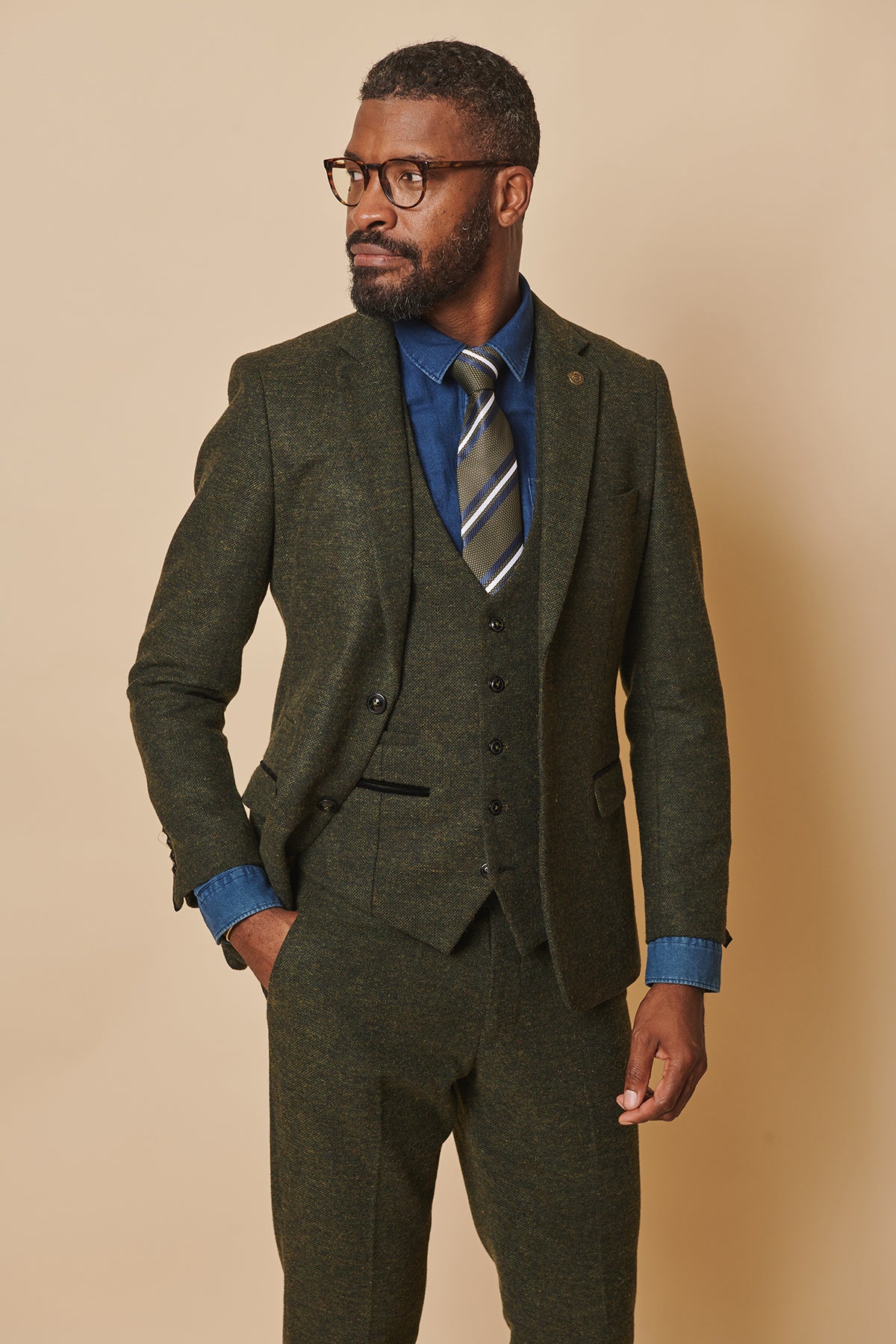 A Black man with glasses and a beard wears the marcdarcy MARLOW Olive Green Tweed Blazer as part of a three-piece suit, styled with a blue shirt and striped tie, his hand in his pocket.