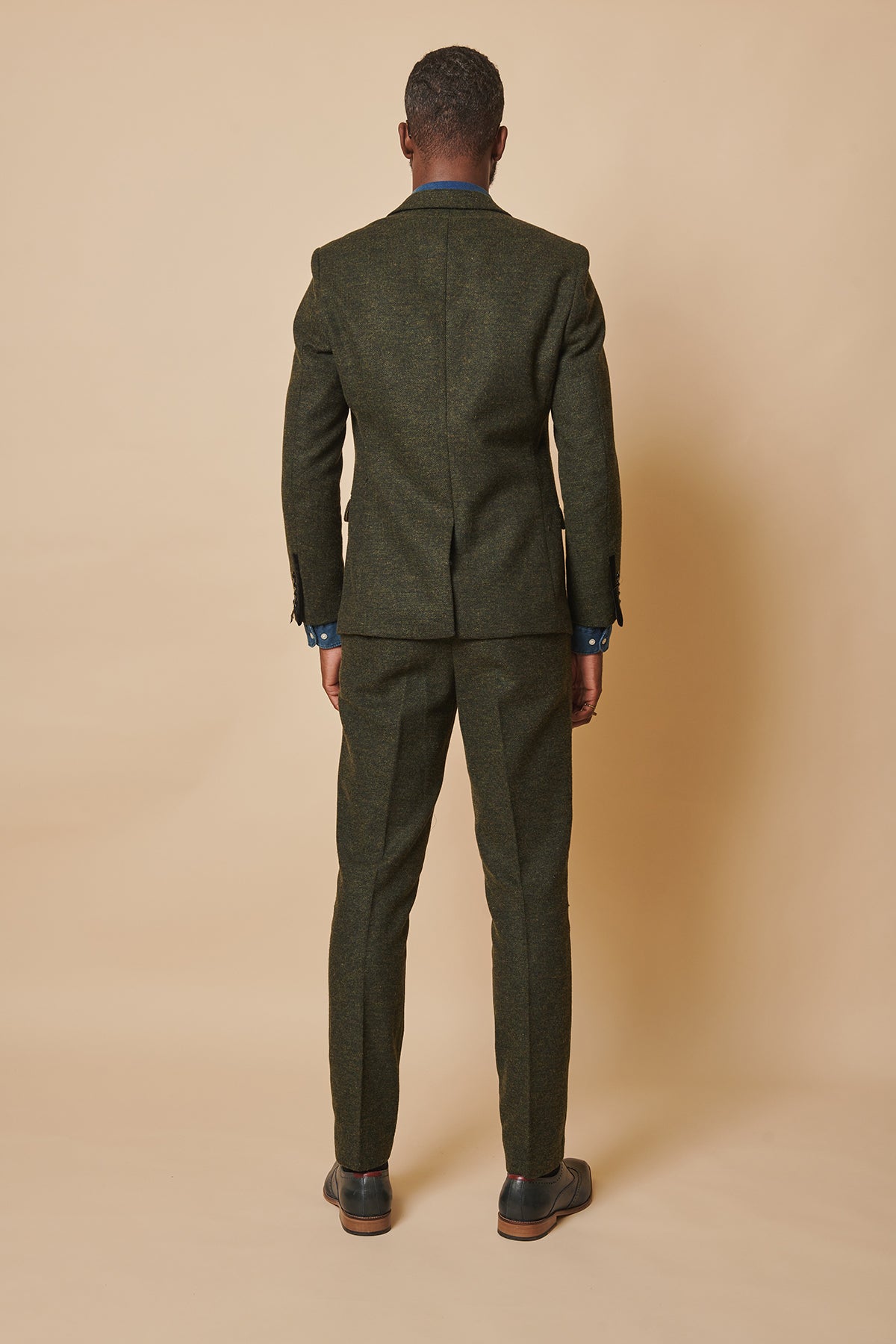 Back view of a man in the MARLOW - Olive Green Tweed Three Piece Suit by marcdarcy, paired with a blue collared shirt and black dress shoes, set against a tan background.