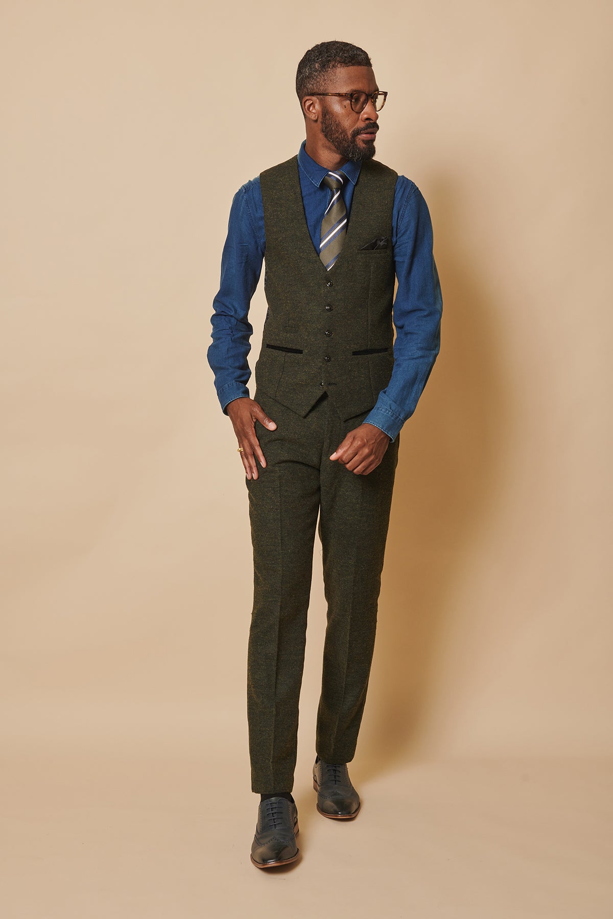 A man in glasses and a formal outfit featuring the marcdarcy MARLOW Olive Green Tweed Three Piece Suit, with vest and pants, blue shirt, striped tie, and black shoes stands against a beige background looking left.