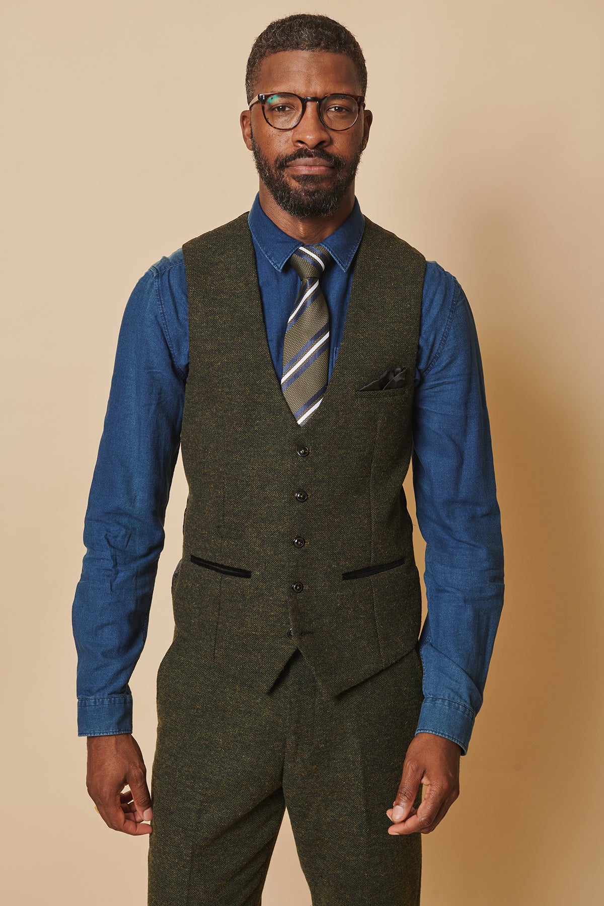 A man with short hair, a beard, and glasses stands against a beige background, wearing a blue shirt, striped tie, and the marcdarcy MARLOW Olive Green Tweed Single Breasted Waistcoat with matching trousers.
