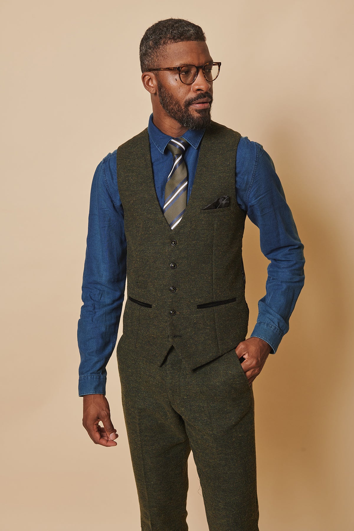 A man in glasses, a blue dress shirt, striped tie, and the marcdarcy MARLOW Olive Green Tweed Single Breasted Waistcoat with matching trousers stands against a beige background, looking to the side with one hand in his pocket.