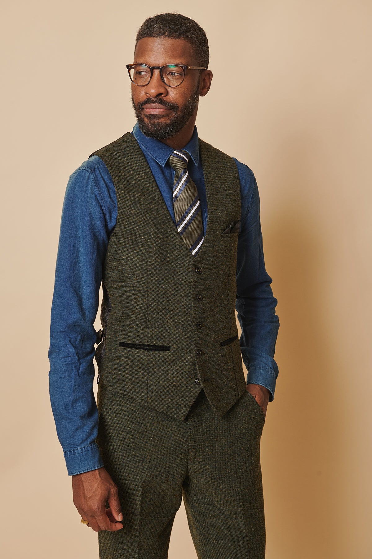 A man with short hair, beard, and glasses wears the MARLOW Olive Green Tweed Single Breasted Waistcoat by marcdarcy with matching trousers, a blue dress shirt, and striped tie. He stands hands in pockets against a plain beige background.