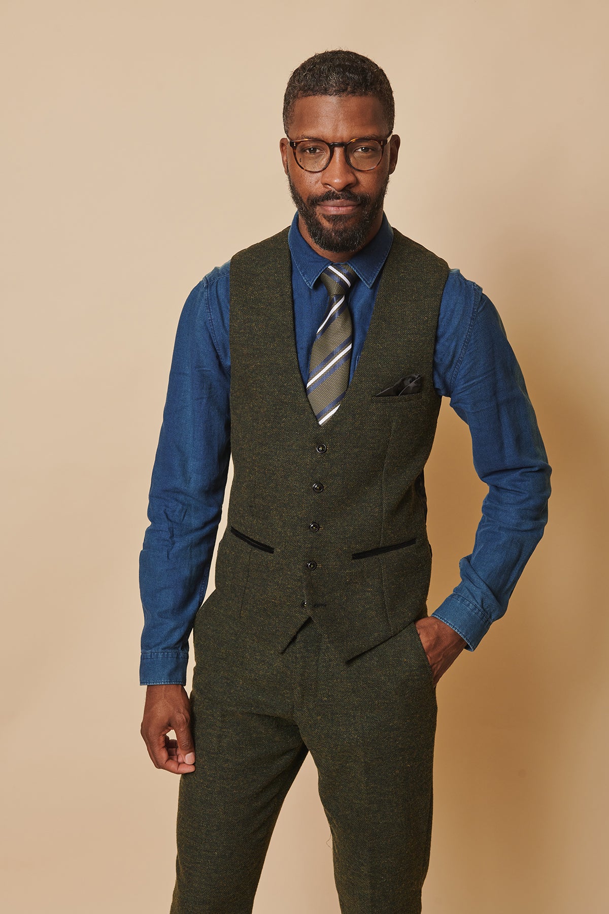 A man with short hair and glasses wears the marcdarcy MARLOW Olive Green Tweed Three Piece Suit, standing confidently against a beige background with one hand in his pocket.