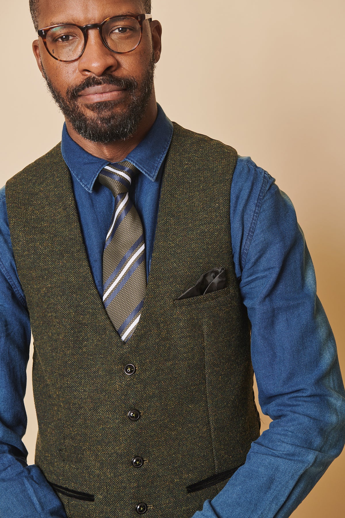 A bearded man with glasses wears a blue denim shirt, striped tie, and the marcdarcy MARLOW Olive Green Tweed Single Breasted Waistcoat with a pocket square, standing against a beige background.