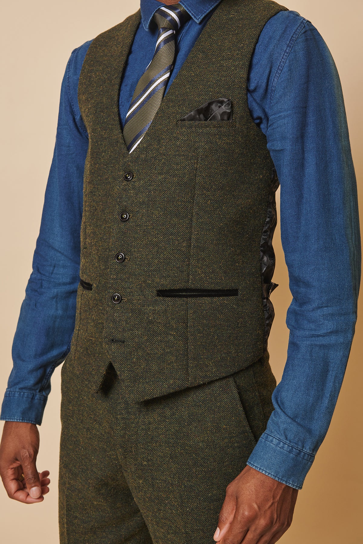 A man wears the marcdarcy MARLOW Olive Green Tweed Single Breasted Waistcoat with matching trousers, a blue denim shirt, striped tie, and black pocket square. The close-up shows the textured waistcoat in a relaxed pose.