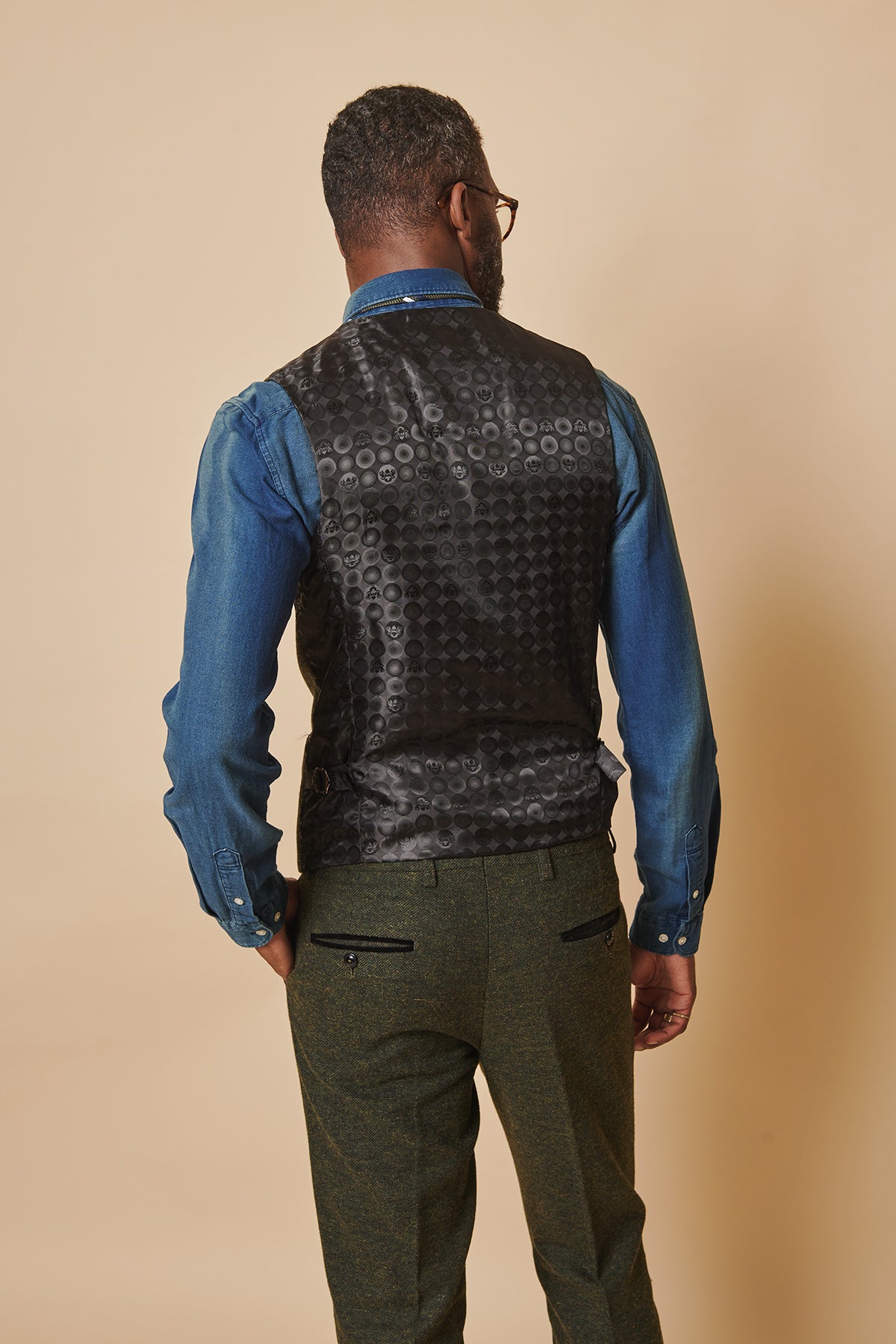 A man stands facing away in a blue denim shirt, marcdarcy’s MARLOW olive green tweed single breasted waistcoat, and green trousers with zippered back pockets, set against a plain beige background.