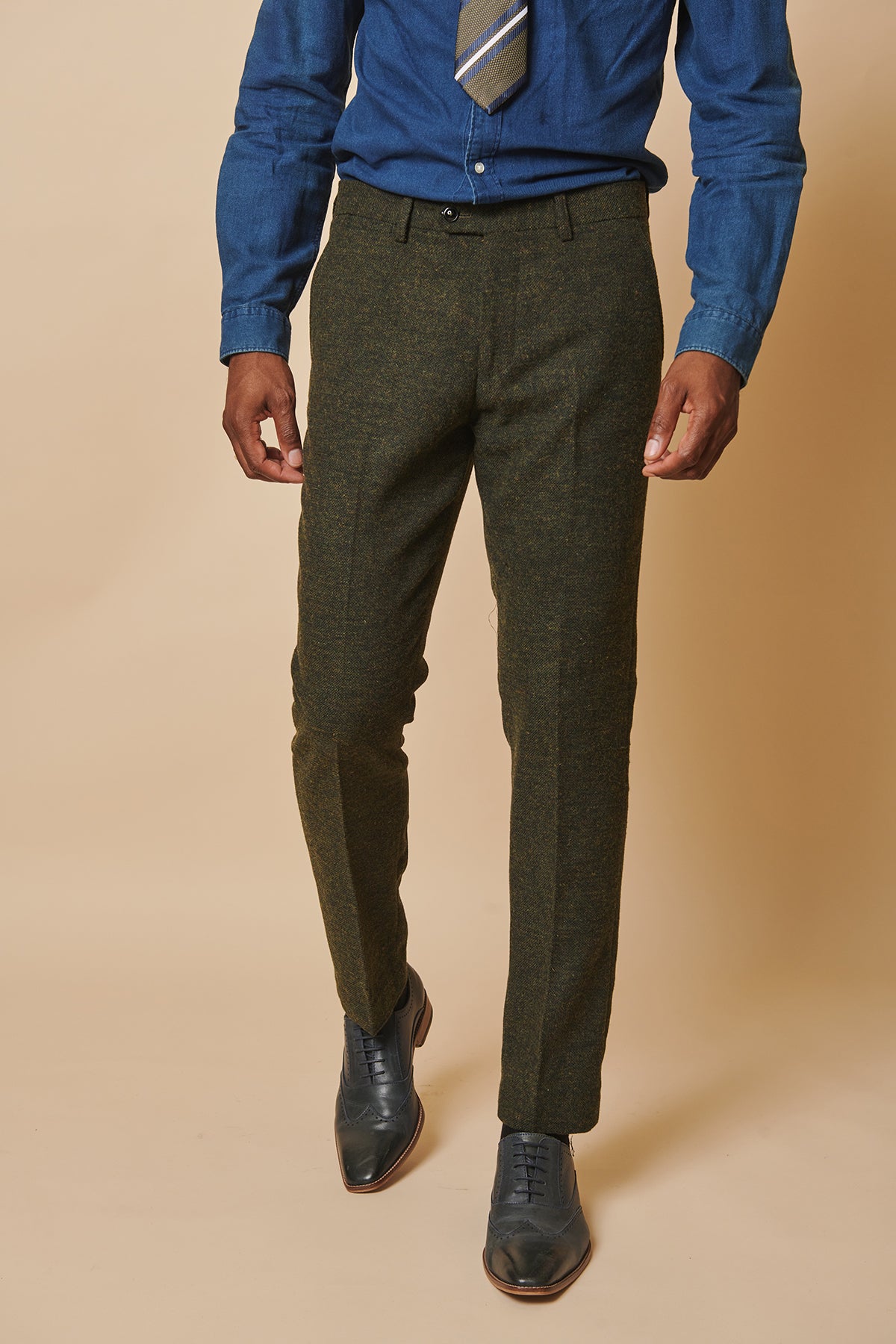 A man in the MARLOW Olive Green Tweed Three Piece Suit by marcdarcy, paired with a blue dress shirt, patterned tie, and dark dress shoes, stands against a beige background.