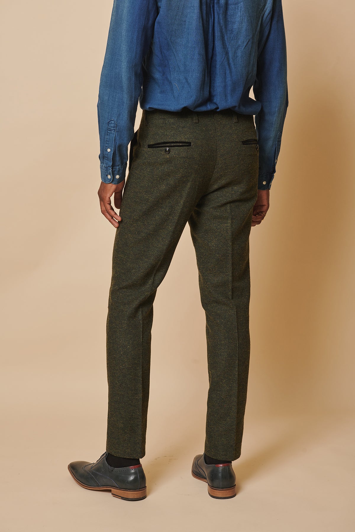 A person stands facing away, wearing a blue button-up shirt, marcdarcys MARLOW Olive Green Tweed Trousers in a slim fit, and dark dress shoes against a beige background.