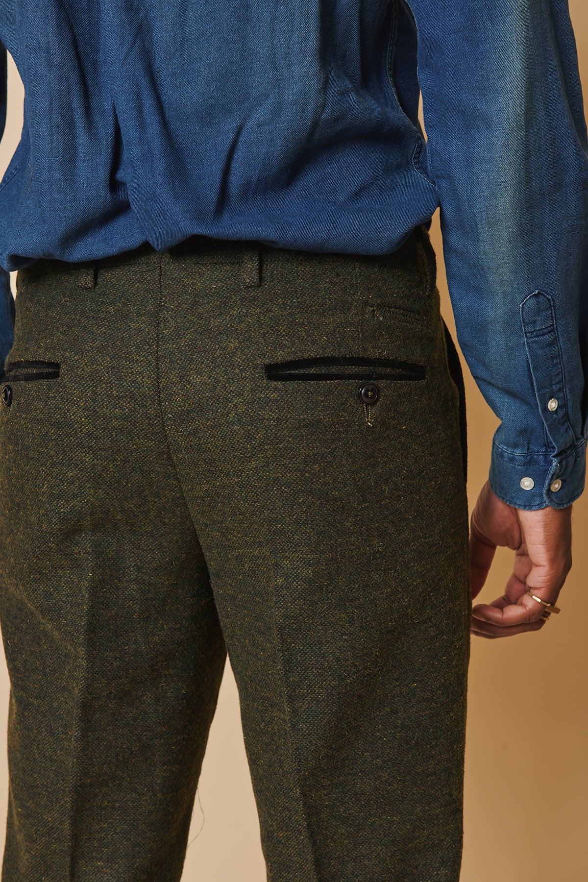 A person shown from the waist down and back view wears a blue long-sleeve shirt with marcdarcy MARLOW Olive Green Tweed Trousers featuring a vintage style, wool blend fabric, back pockets, and black button details.