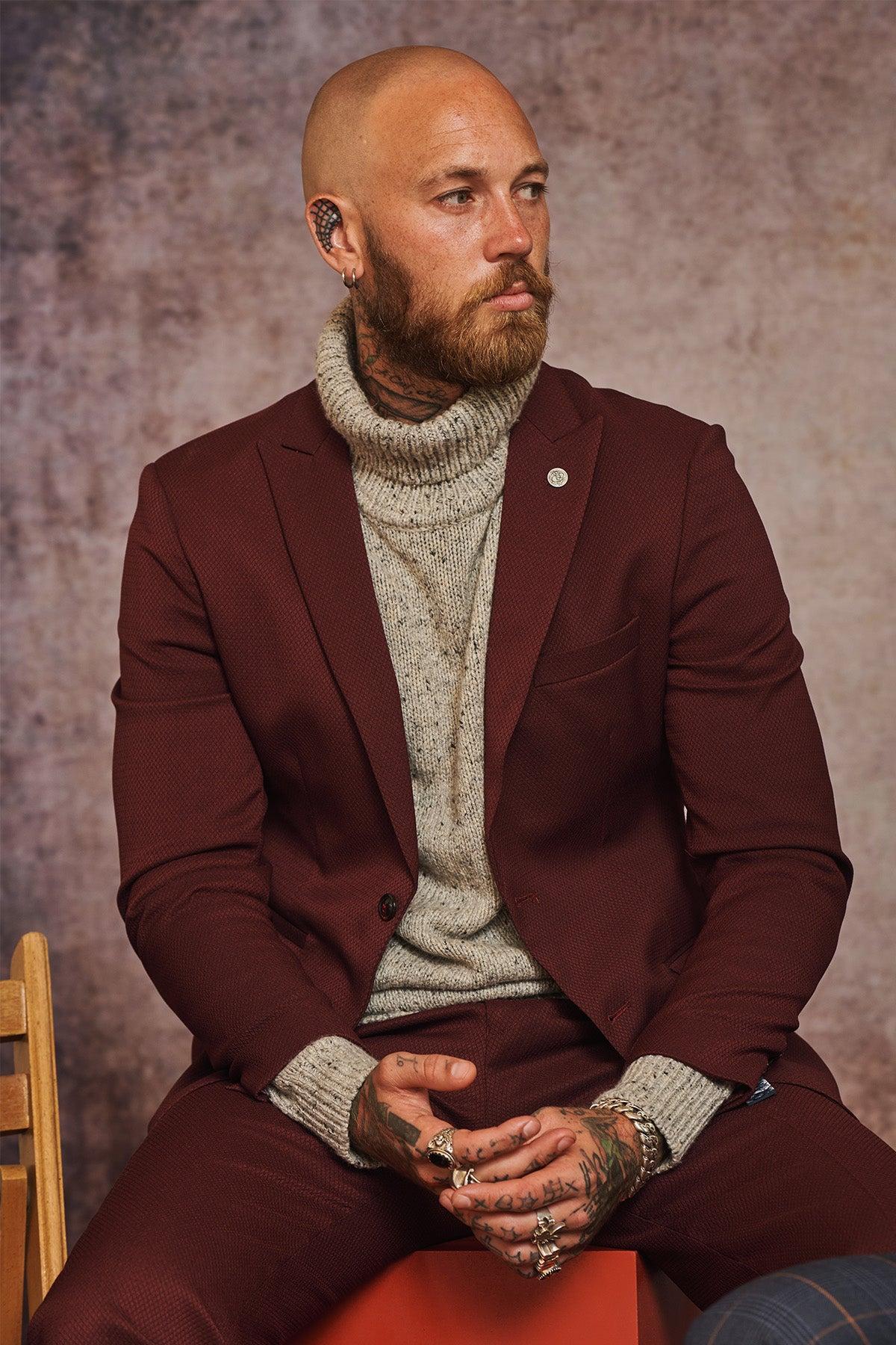 A bald, bearded man with neck and hand tattoos wears the marcdarcy MAX Wine Two Piece Suit over a beige turtleneck, finishing his look with a camouflage pocket square.