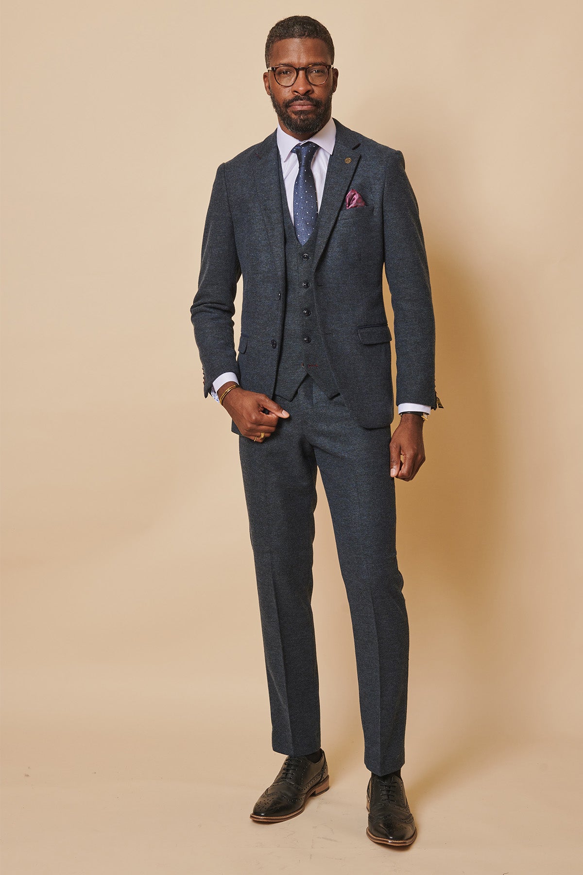 A man stands against a beige background in the marcdarcy MARLOW Blue Tweed Three Piece Suit, styled with a light blue shirt, navy tie, and purple pocket square. He wears glasses and black shoes, looking confidently at the camera.