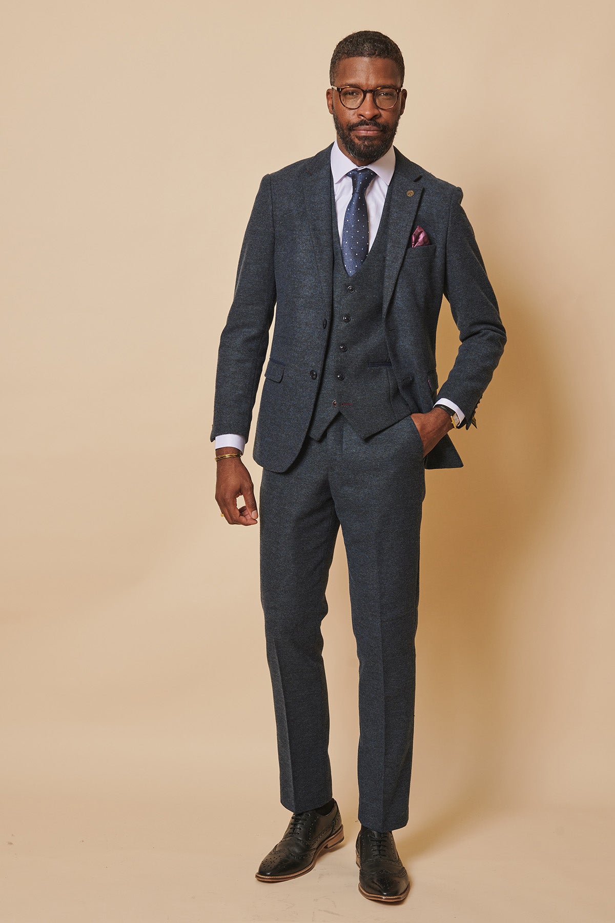 A man stands against a beige background wearing the marcdarcy MARLOW Blue Tweed Three Piece Suit in slim fit, styled with a white shirt, tie, pocket square, glasses, black shoes, and one hand in his pocket.