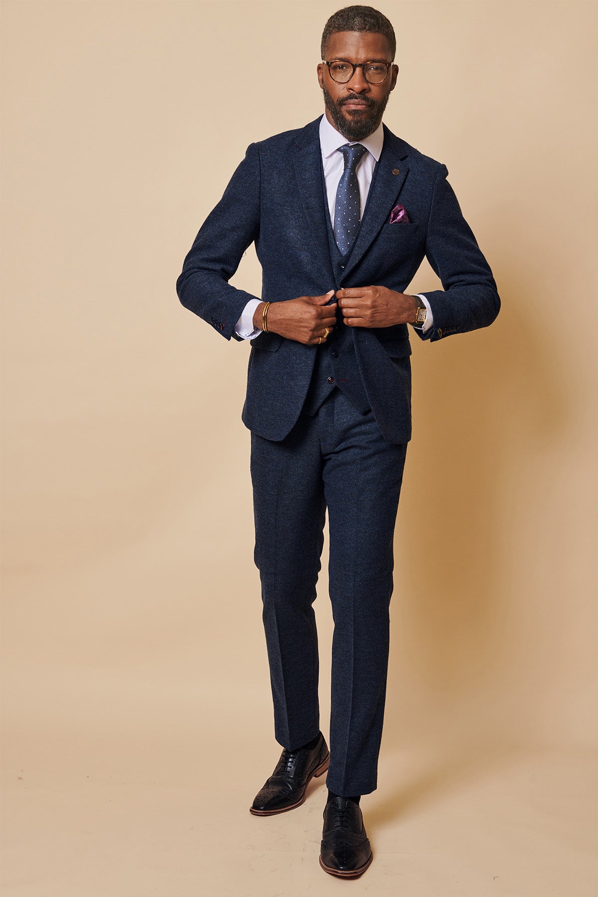 A man in a marcdarcy MARLOW navy tweed three piece suit with circle print lining stands against a beige background, buttoning his jacket. He accessorizes with glasses, a beard, white dress shirt, tie, and dress shoes.