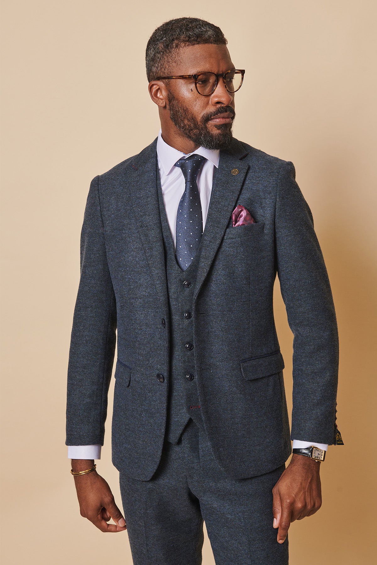 A Black man wearing the marcdarcy MARLOW Blue Tweed Blazer with a white shirt, navy polka dot tie, glasses, and a gold bracelet poses against a tan background.