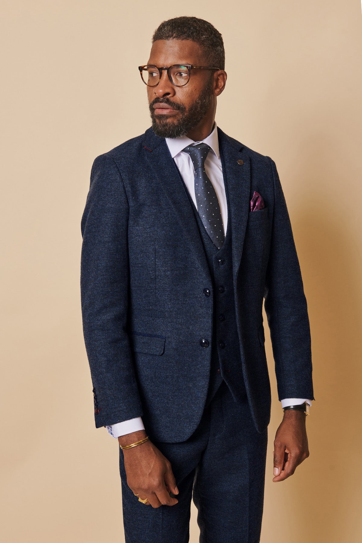 A man wearing glasses and the marcdarcy MARLOW Navy Tweed Blazer with a white shirt, polka dot tie, and purple pocket square stands against a beige background, looking to his right.
