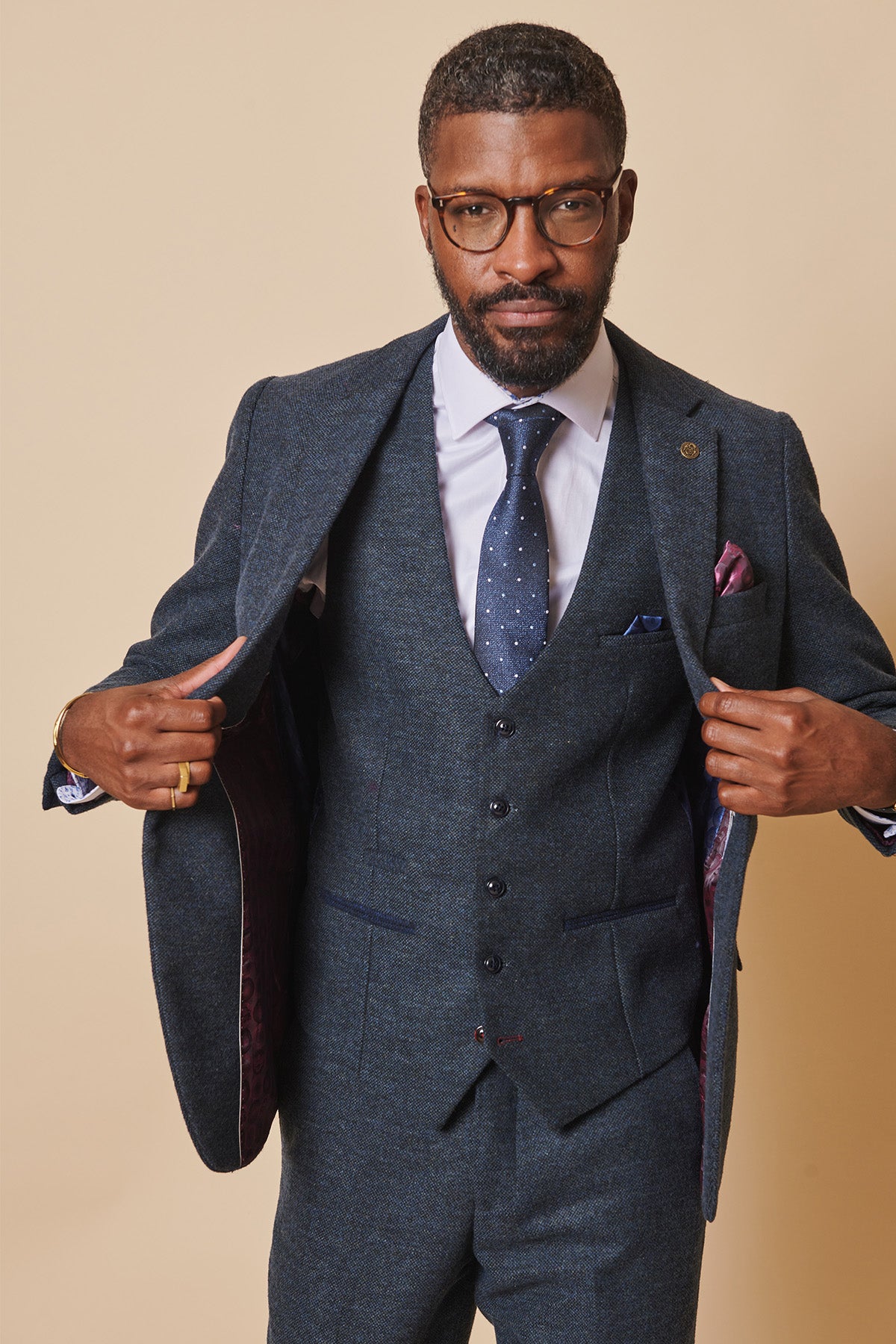 A man with short hair, beard, and glasses models the marcdarcy MARLOW Blue Tweed Three Piece Suit with a white shirt and navy dotted tie. He opens the jacket to reveal the lining, facing the camera against a beige background.