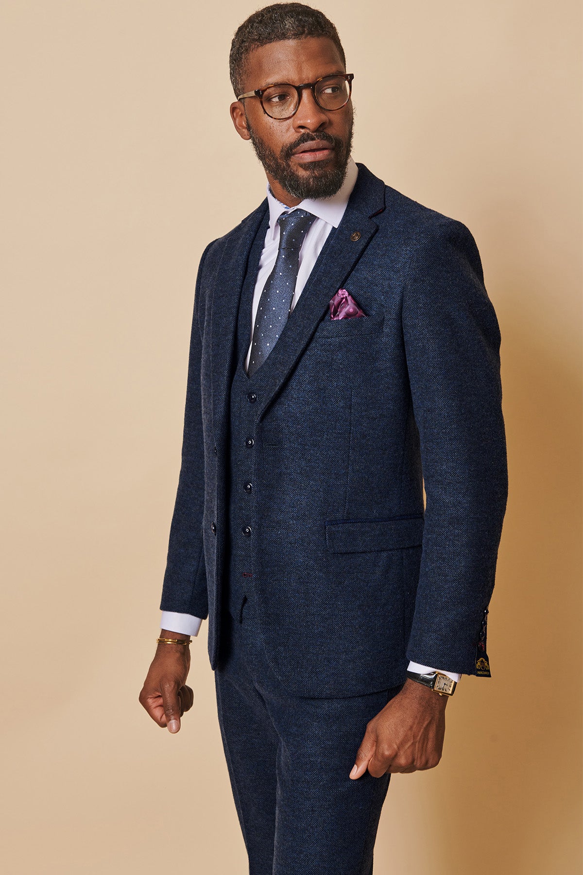 A man with short hair, a beard, and glasses wears the marcdarcy MARLOW Navy Tweed Blazer in a slim fit with a matching vest, white shirt, dotted navy tie, and purple pocket square against a beige background.