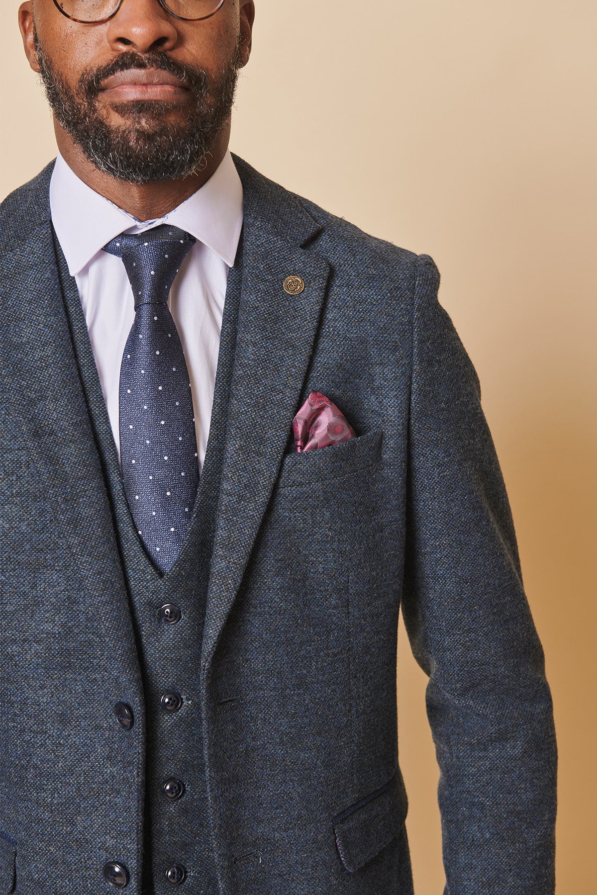A man models the marcdarcy MARLOW Blue Tweed Three Piece Suit with a white shirt, navy polka-dot tie, glasses, and pink pocket square, shown chest up against a beige background.