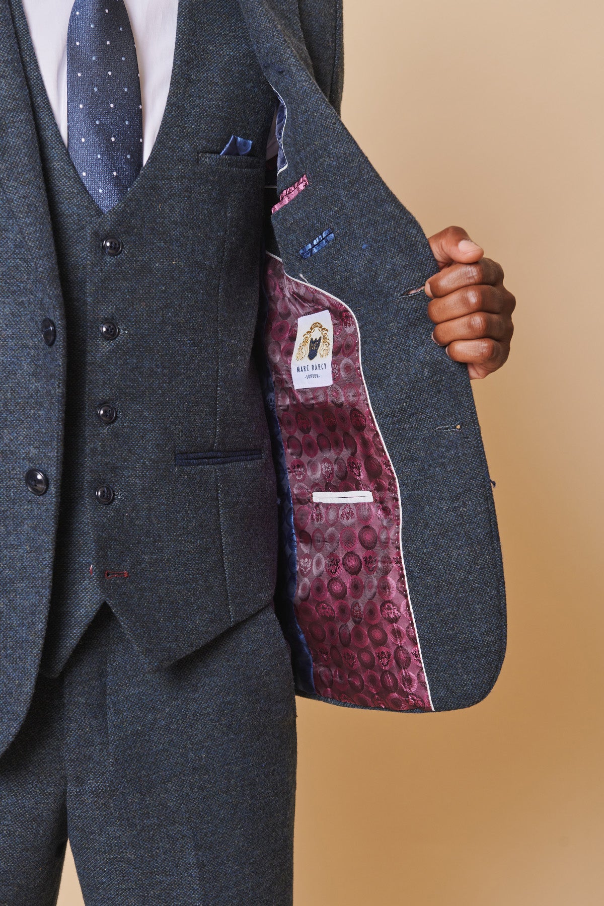 A person in a MARLOW Blue Tweed Blazer by marcdarcy and a blue tie opens their jacket to reveal a purple patterned lining and a “RAYE BEAUX” label, against a beige background.