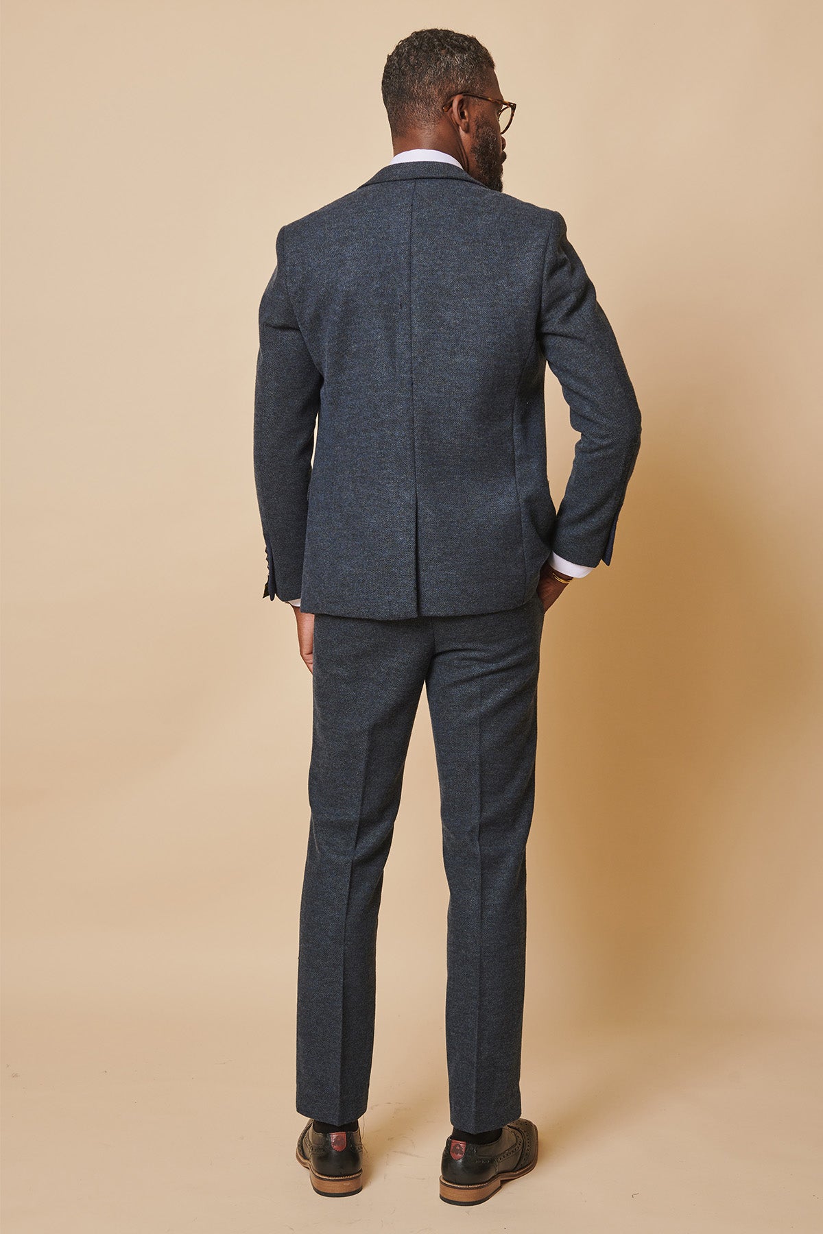 A man shown from behind wears the marcdarcy MARLOW blue tweed three piece suit in slim fit with a white shirt and brown brogues, set against a beige background.